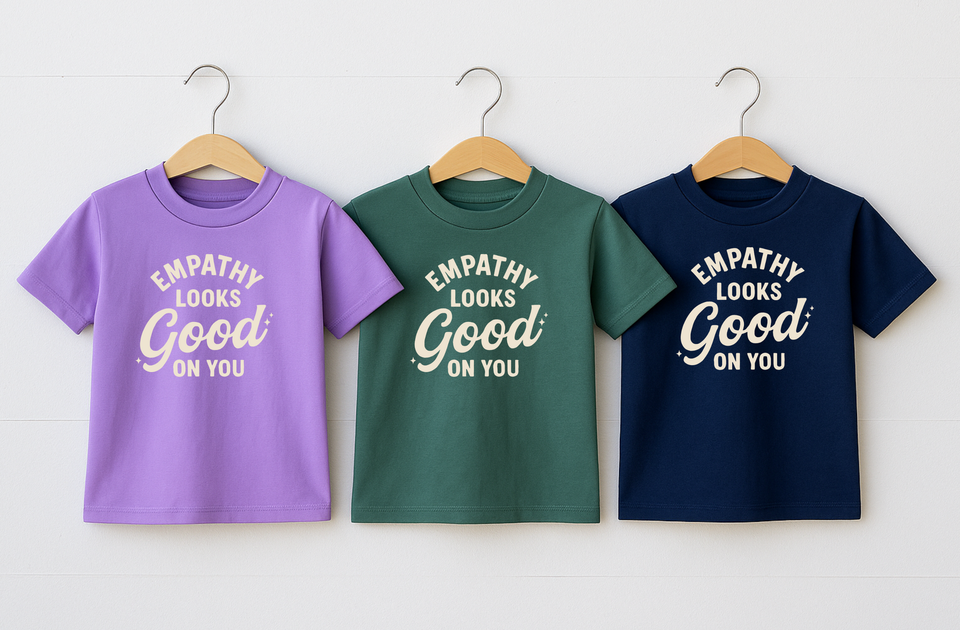 Three t-shirts in purple, green, and blue with 'Empathy Looks Good On You' text on hangers against a white background. empathy tee shirts for kids