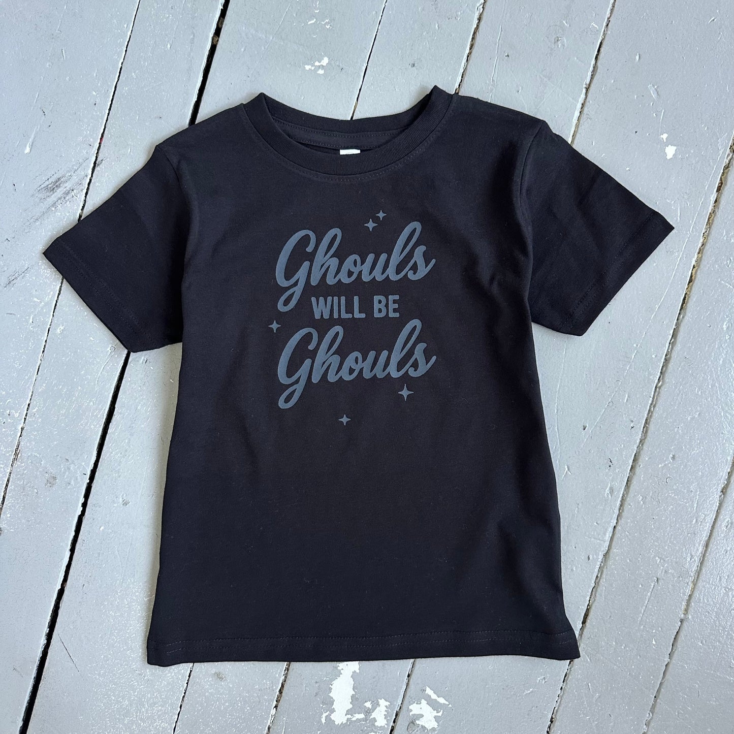 Black t-shirt with 'Ghouls Will Be Ghouls' text on a light wooden floor.