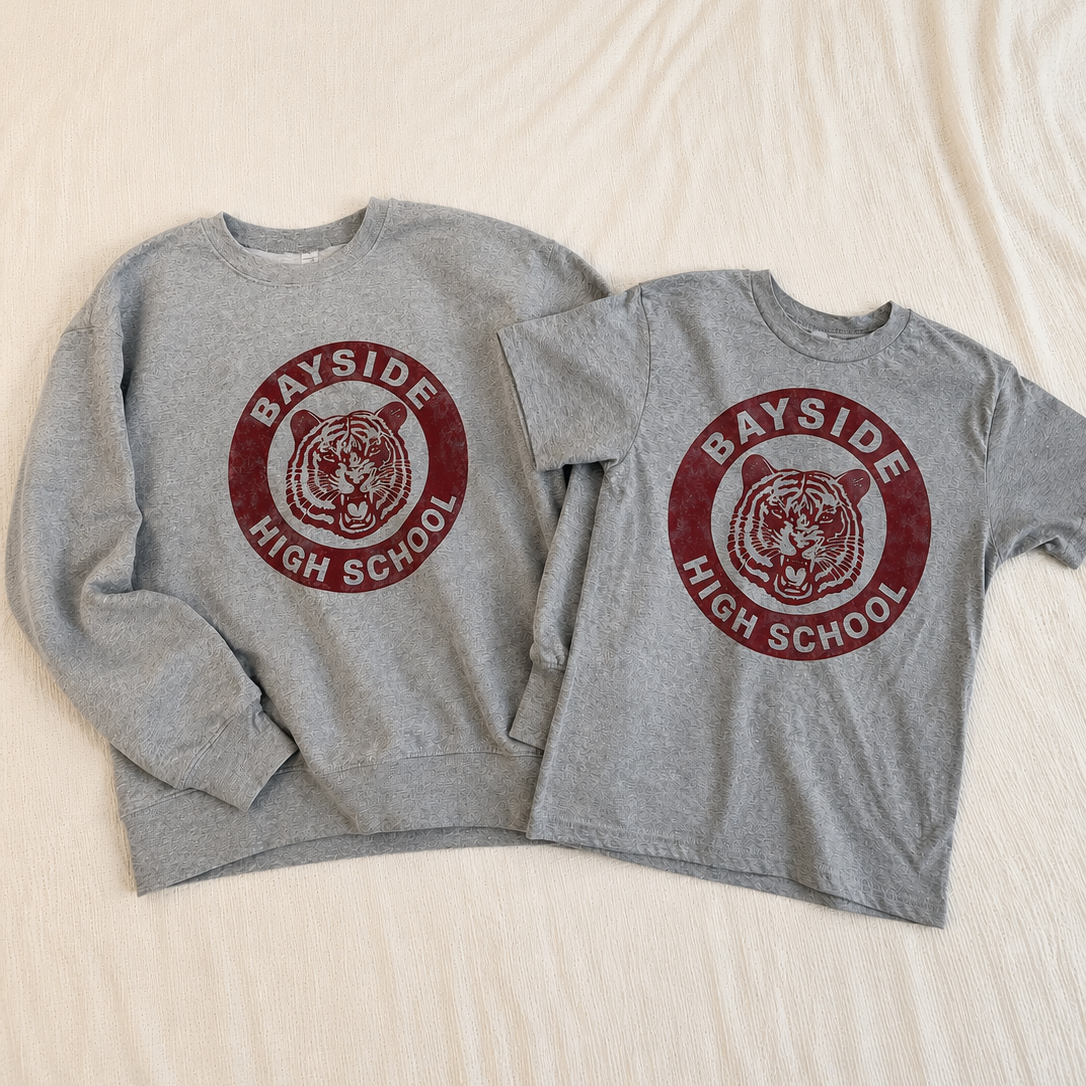 Two heather gray crewneck sweatshirts laid flat on a light beige fabric background. Both sweatshirts feature a circular red graphic with a tiger head and the text 'BAYSIDE HIGH SCHOOL'.