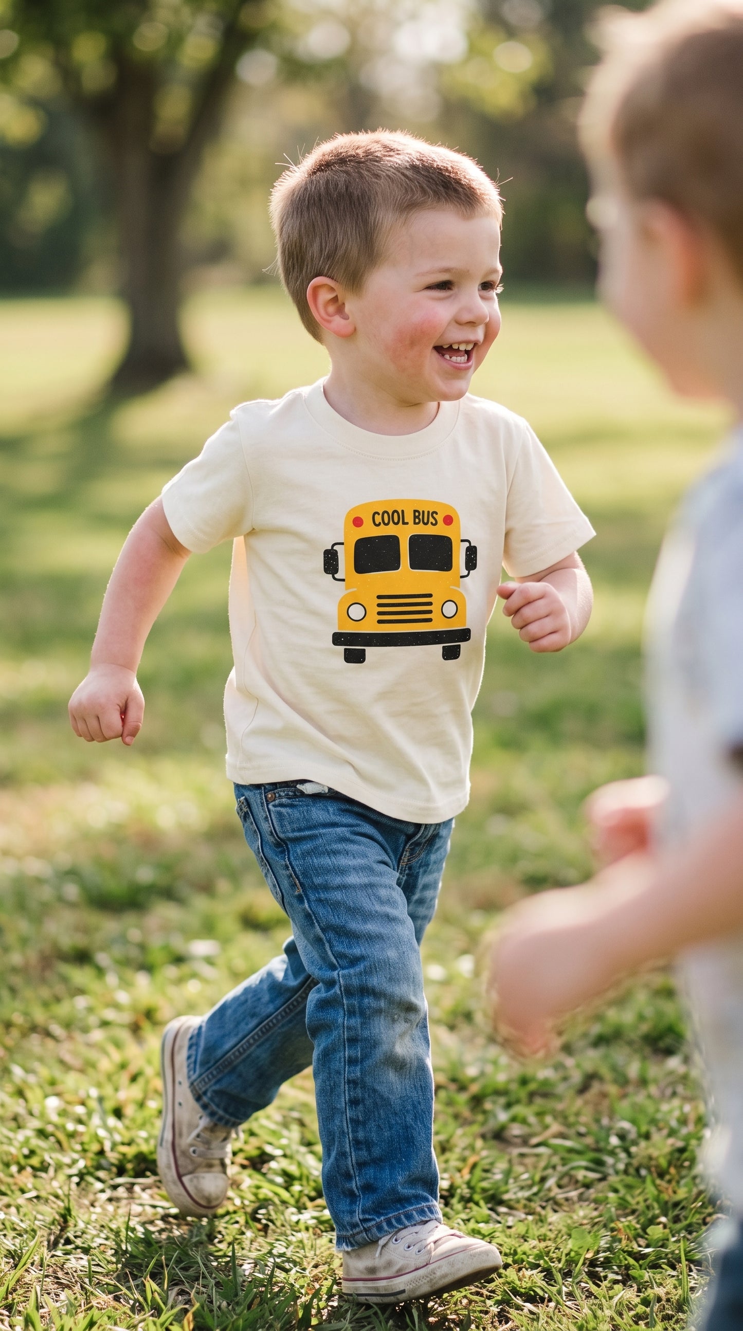 A young boy running outdoors on a grassy lawn wearing a cream-colored t-shirt with a yellow school bus graphic and blue jeans.