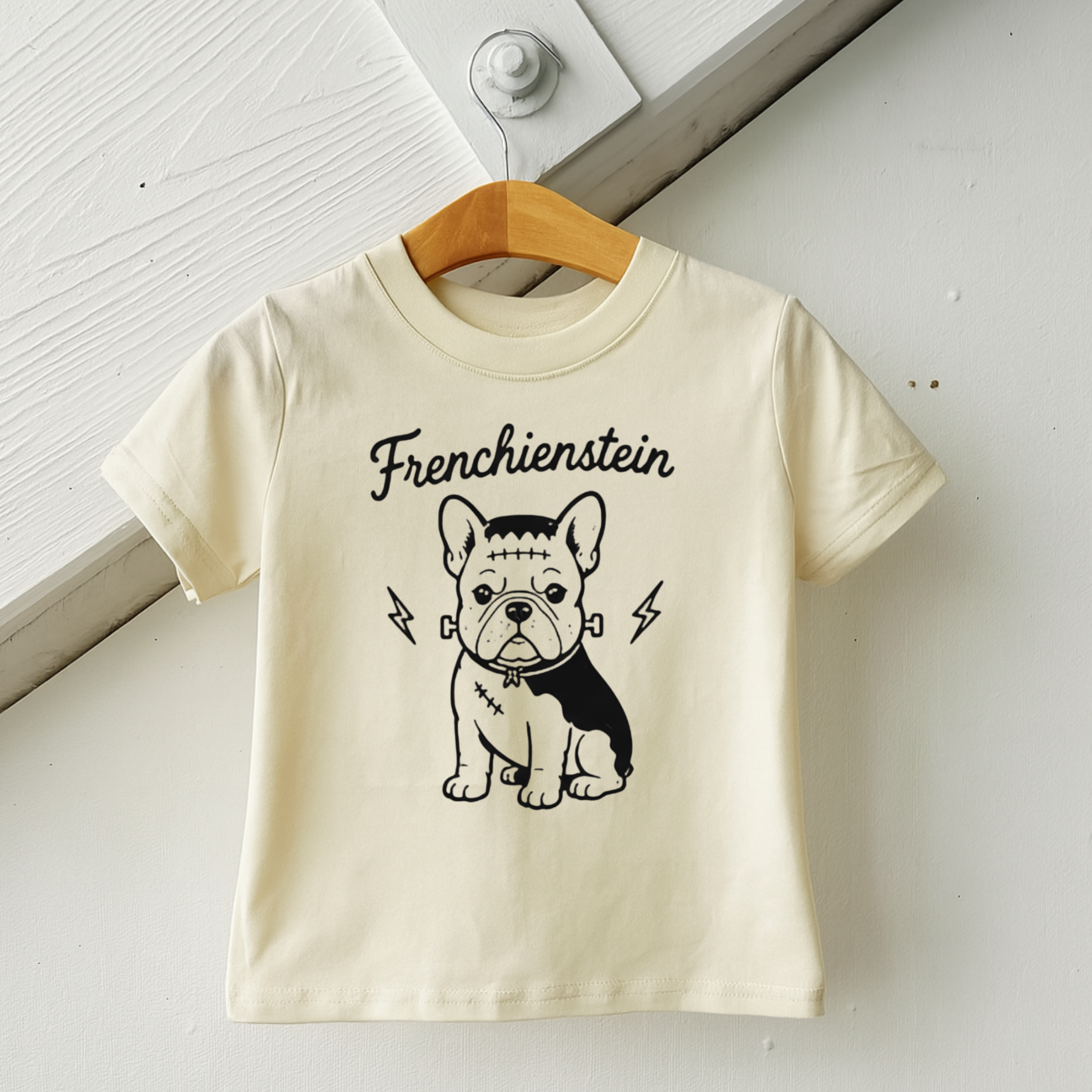 A cream-colored short-sleeve t-shirt hanging on a wooden hanger against a white paneled wall. The shirt features a black graphic of a French Bulldog with the word 'Frenchienstein' printed above it.