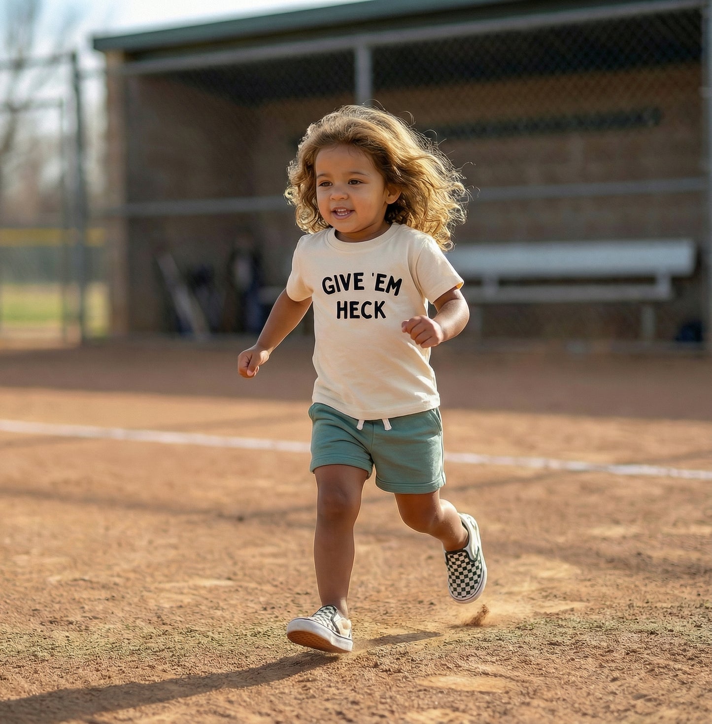 Child running on a baseball field wearing a 'GIVE EM HECK' shirt. 
