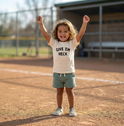 Child on a baseball field wearing a 'Give Em' Heck' t-shirt with arms raised. funny baseball shirt for kids
