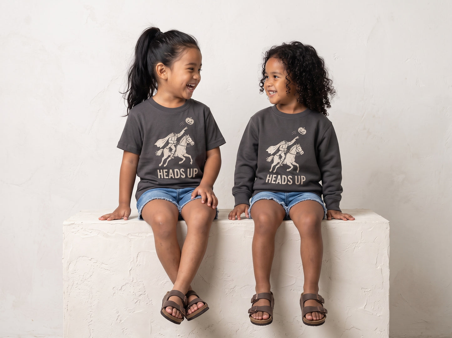 Two young girls sitting side-by-side on a white textured block, wearing matching dark gray crewneck sweatshirts with a white graphic of a cowboy on a horse and the text 'HEADS UP'.