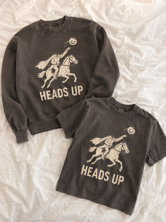 Two matching dark gray crewneck sweatshirts laid flat on a white wrinkled fabric background. The larger sweatshirt features a graphic of a knight on a rearing horse with a glowing pumpkin headlight, and the text 'HEADS UP'.