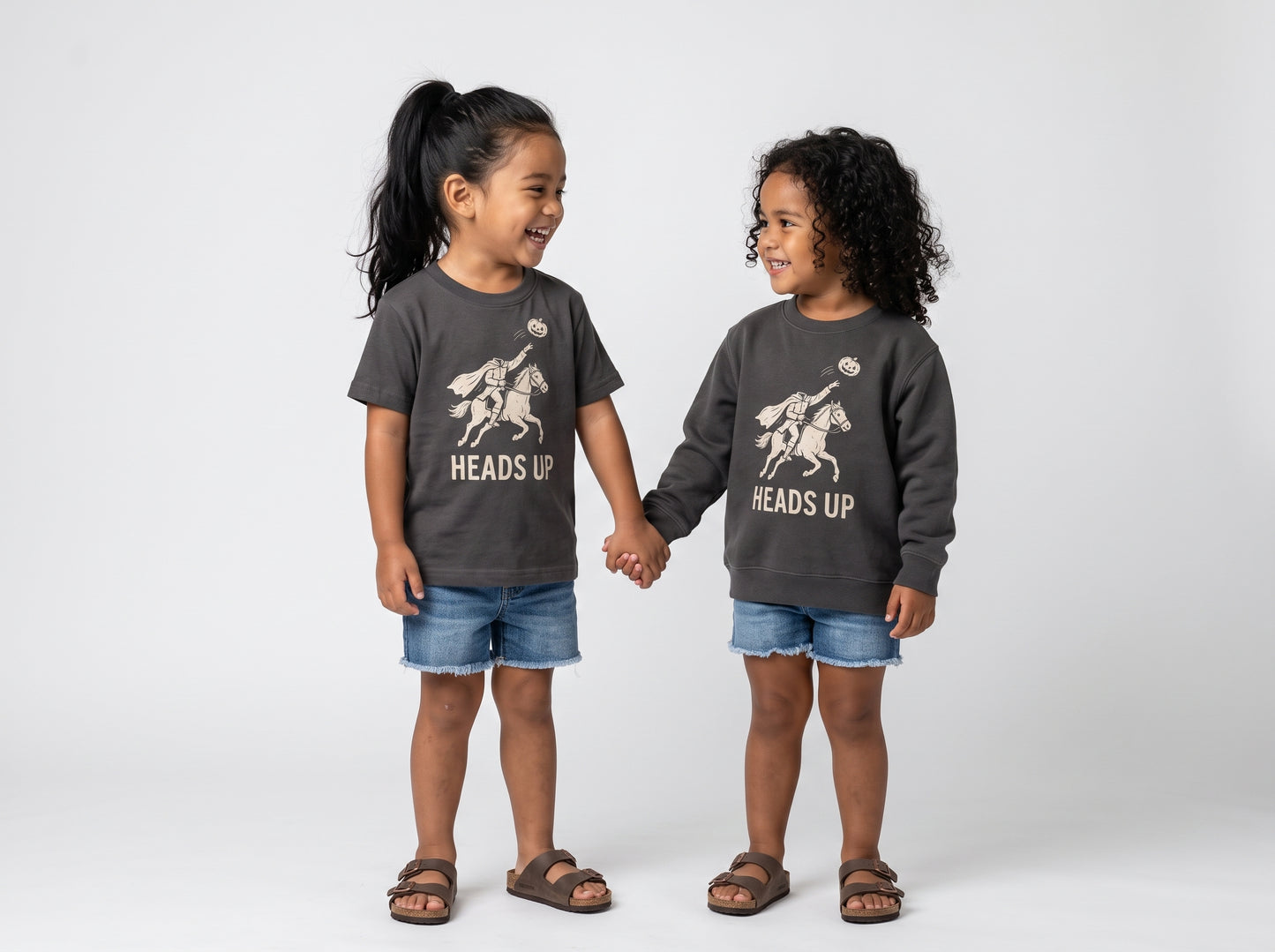 Two young girls standing and holding hands, wearing matching dark gray crewneck sweatshirts with a graphic of a knight on a horse and the text 'HEADS UP', paired with denim shorts and brown sandals, against a light gray studio background.