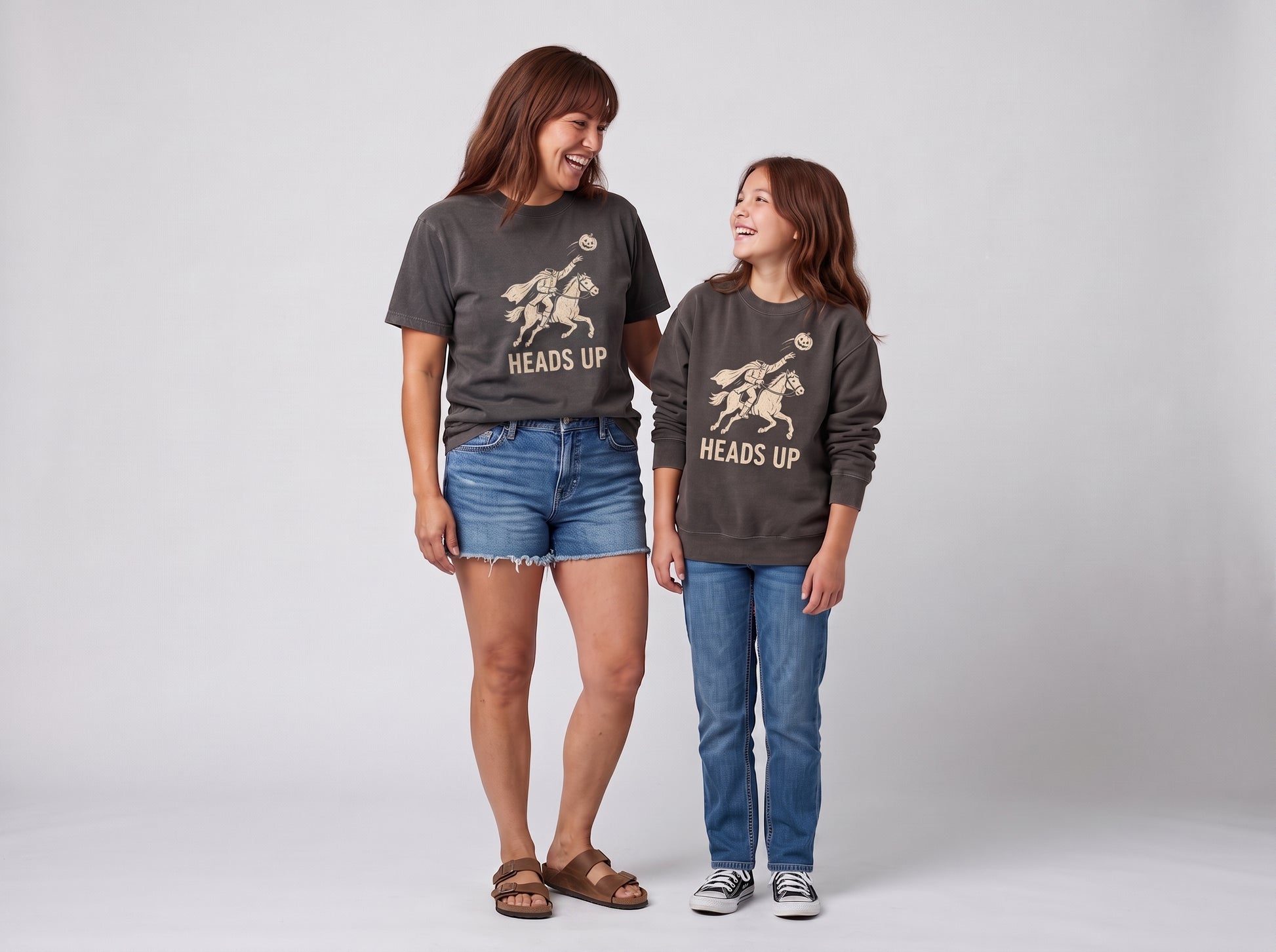 An adult woman and a young girl standing side by side in matching dark gray graphic t-shirts with a gold horse design and the text 'HEADS UP'. The woman wears denim shorts and sandals, while the girl wears blue jeans and sneakers.
