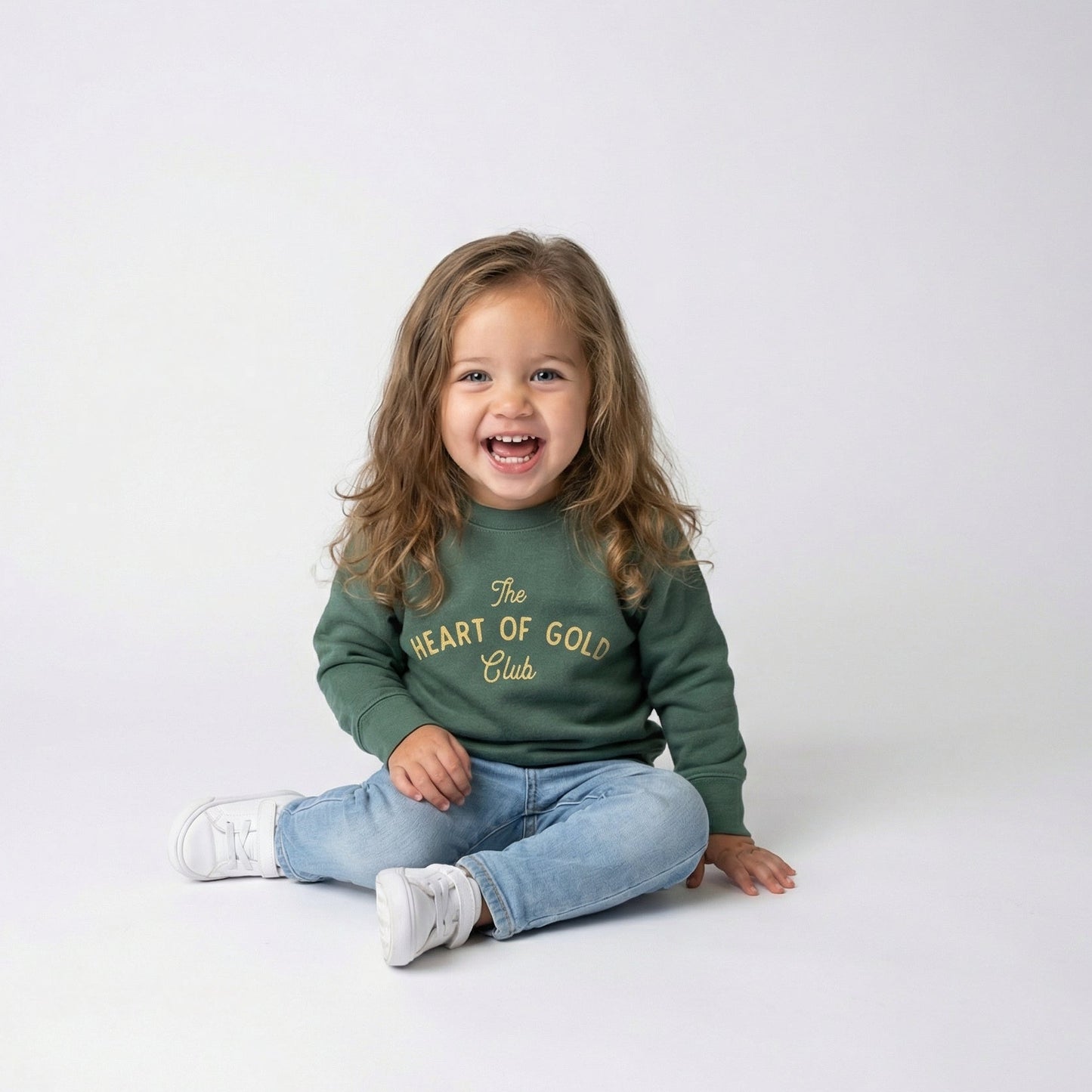 Child wearing a green heart of gold st patty sweatshirt with text, sitting on a white floor.