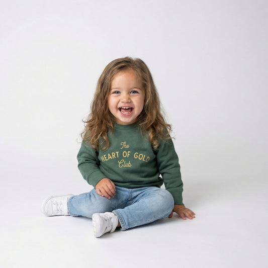 Child wearing a green heart of gold st patty sweatshirt with text, sitting on a white floor.