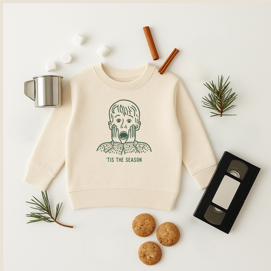 Beige sweatshirt with graphic design on a white surface with holiday items. tis the season Kevin illustration home alone
