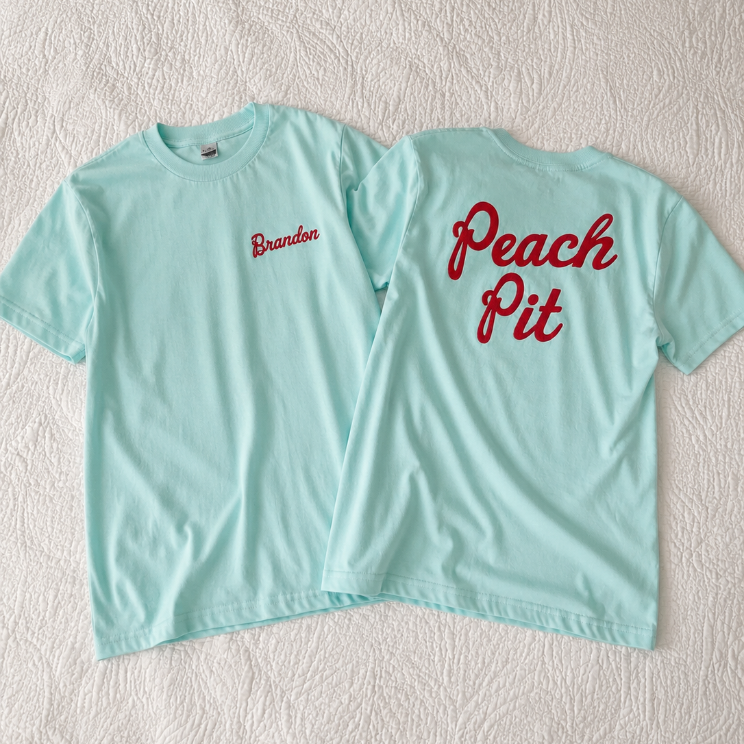 Two mint green t-shirts laid flat on a textured white fabric background, showing the front and back. The front has 'Brandbo' embroidered in red on the left chest, and the back has 'Peach Pit' embroidered in red script.