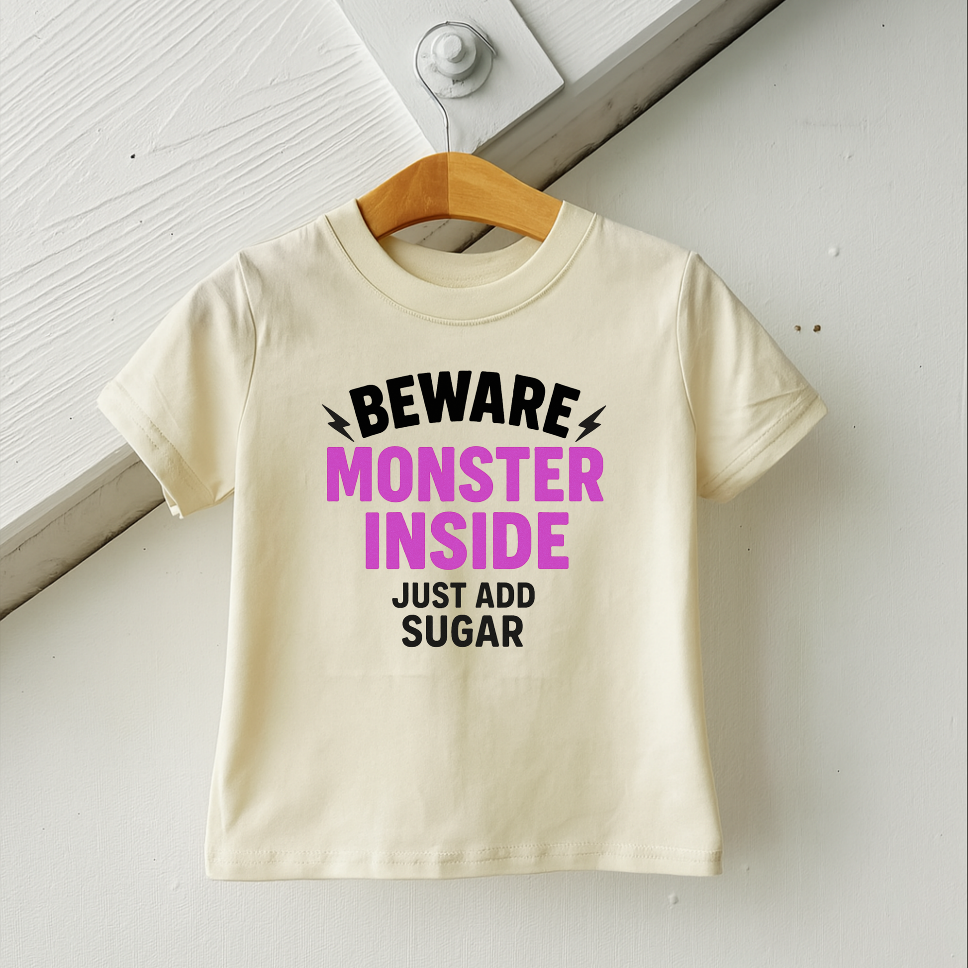 Beige t-shirt with humorous text on a wooden hanger against a white wall. trick or treat toddler tee halloween shirt for girls beware monster inside 
