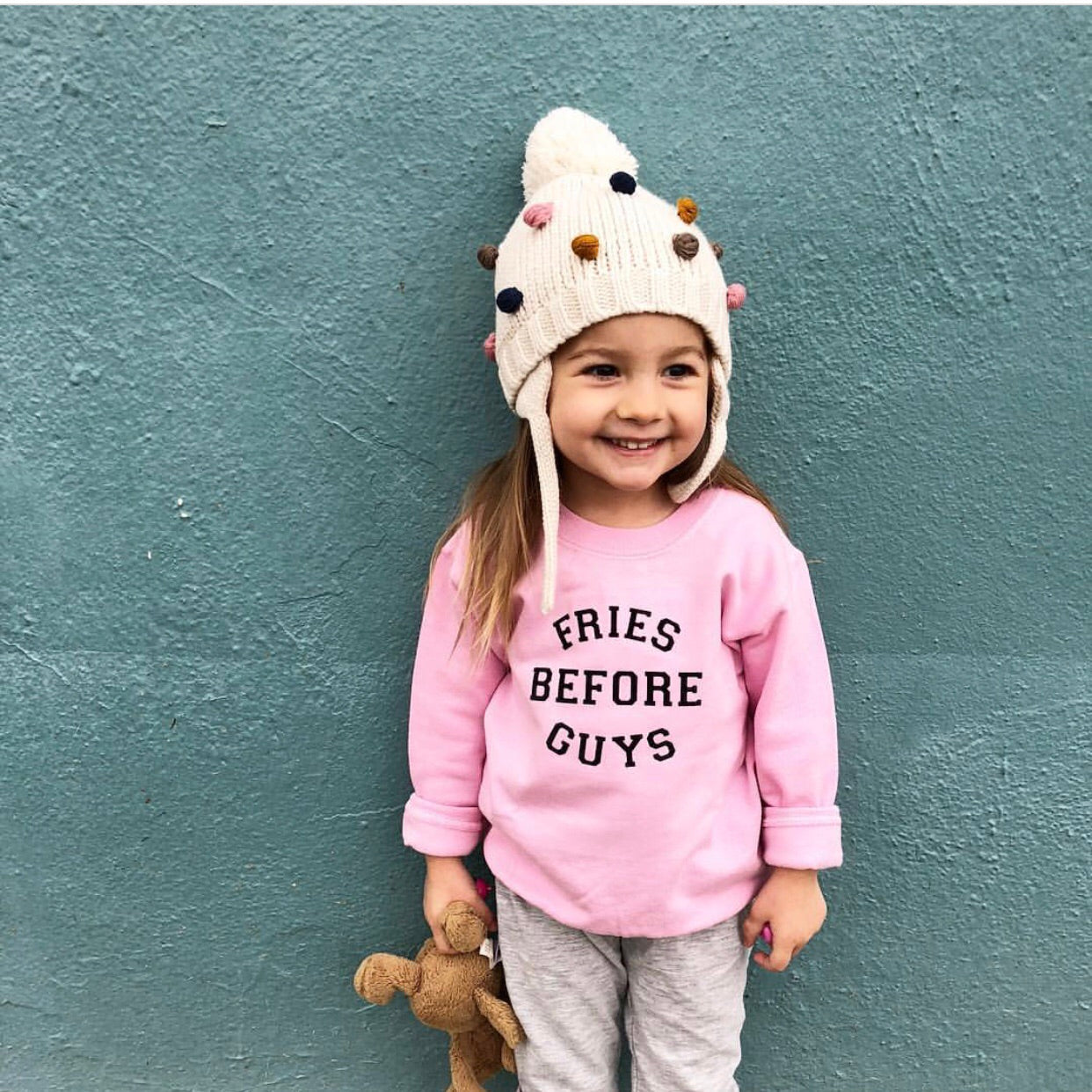 girl standing infront of green wall wearing a winter hat and friens before guys sweatshirt carrying teddy bear